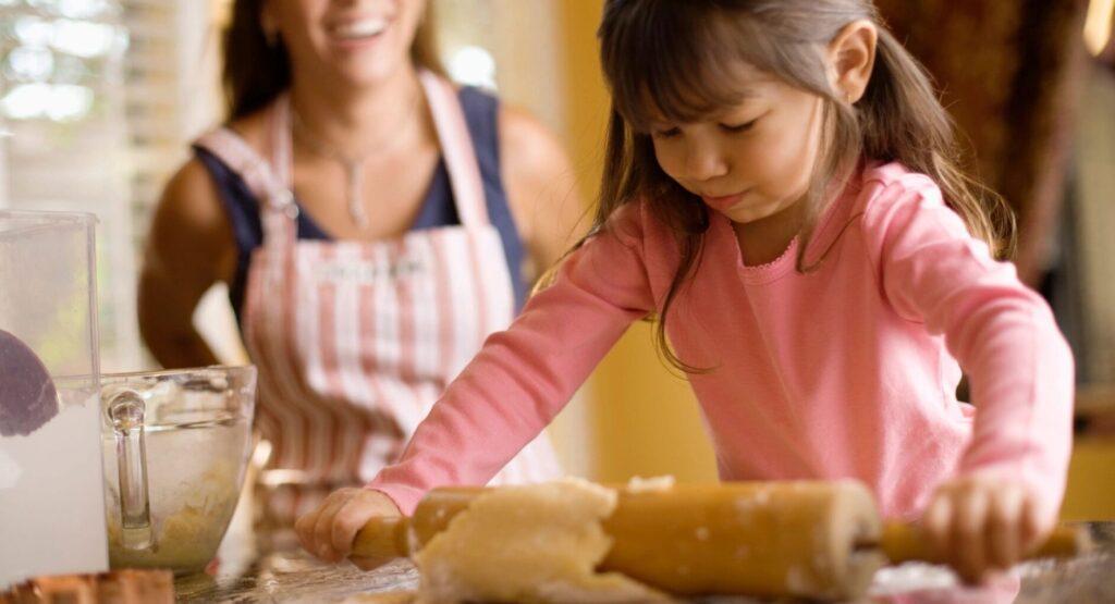 kid using a rolling pin to rollout out pastry with an adult laughing behind her